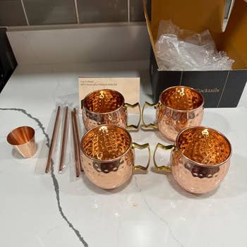 Four copper mugs with handles and a measuring cup, alongside straws on a kitchen counter, possibly for Moscow Mule cocktails