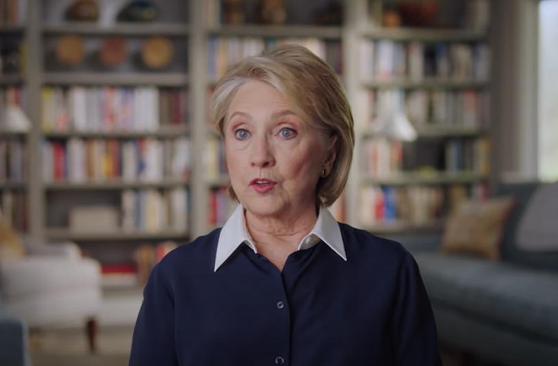 Hillary Clinton in a dark blouse with a collar, sitting, with a bookshelf in the background