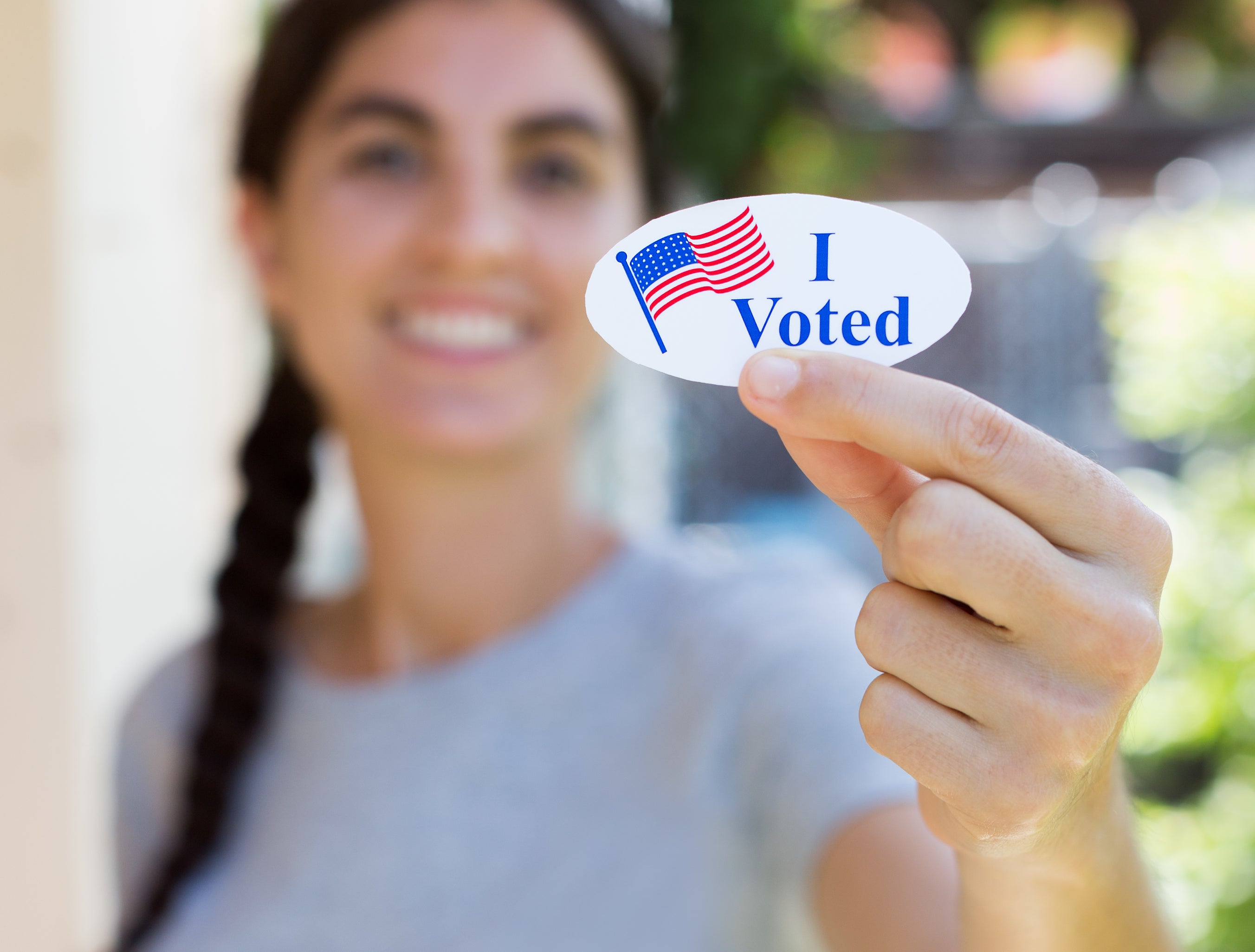A woman holding up an &quot;I Voted&quot; sticker, smiling