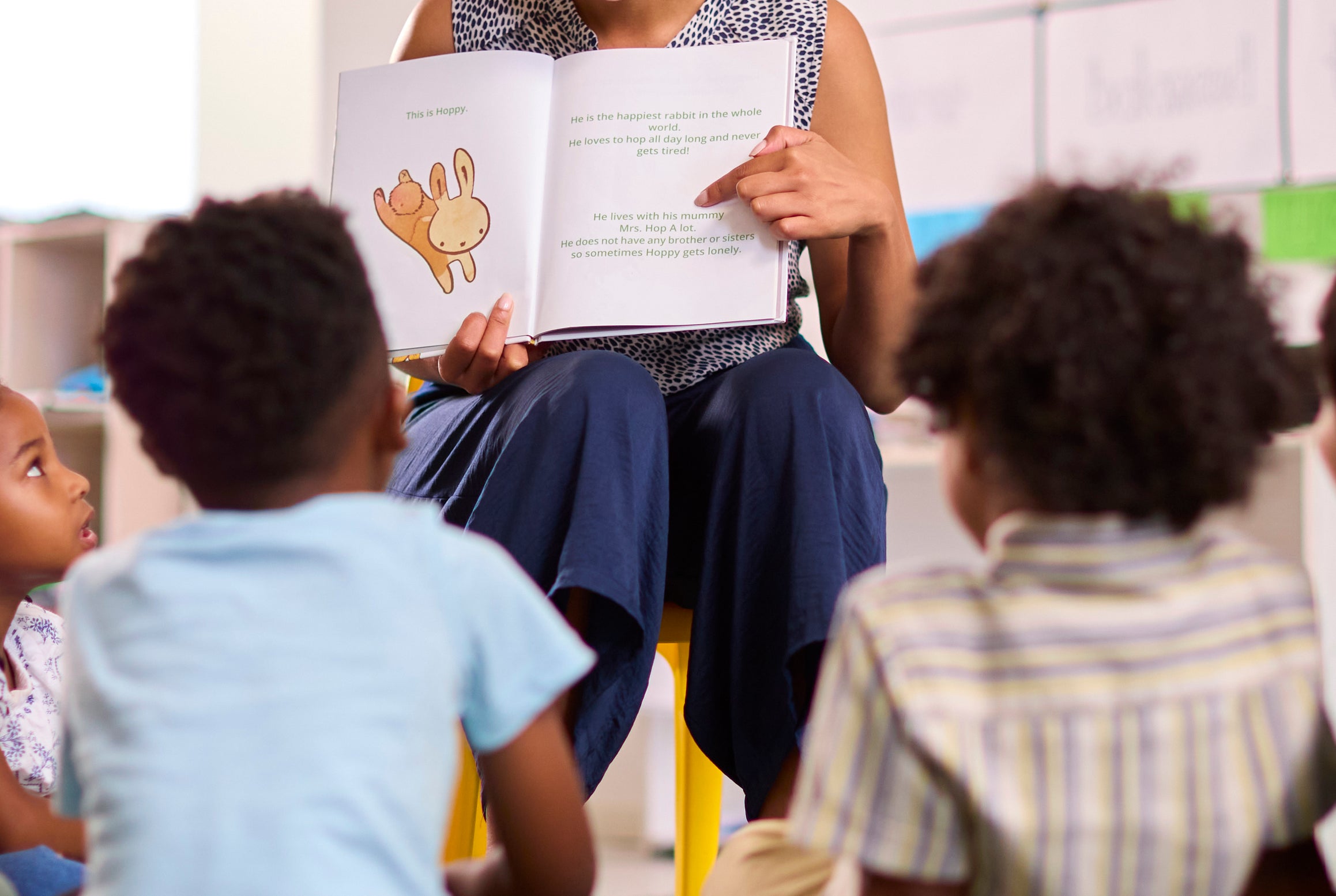 Teacher reads to children sitting on floor, pointing to a book, in a cheerful classroom setting