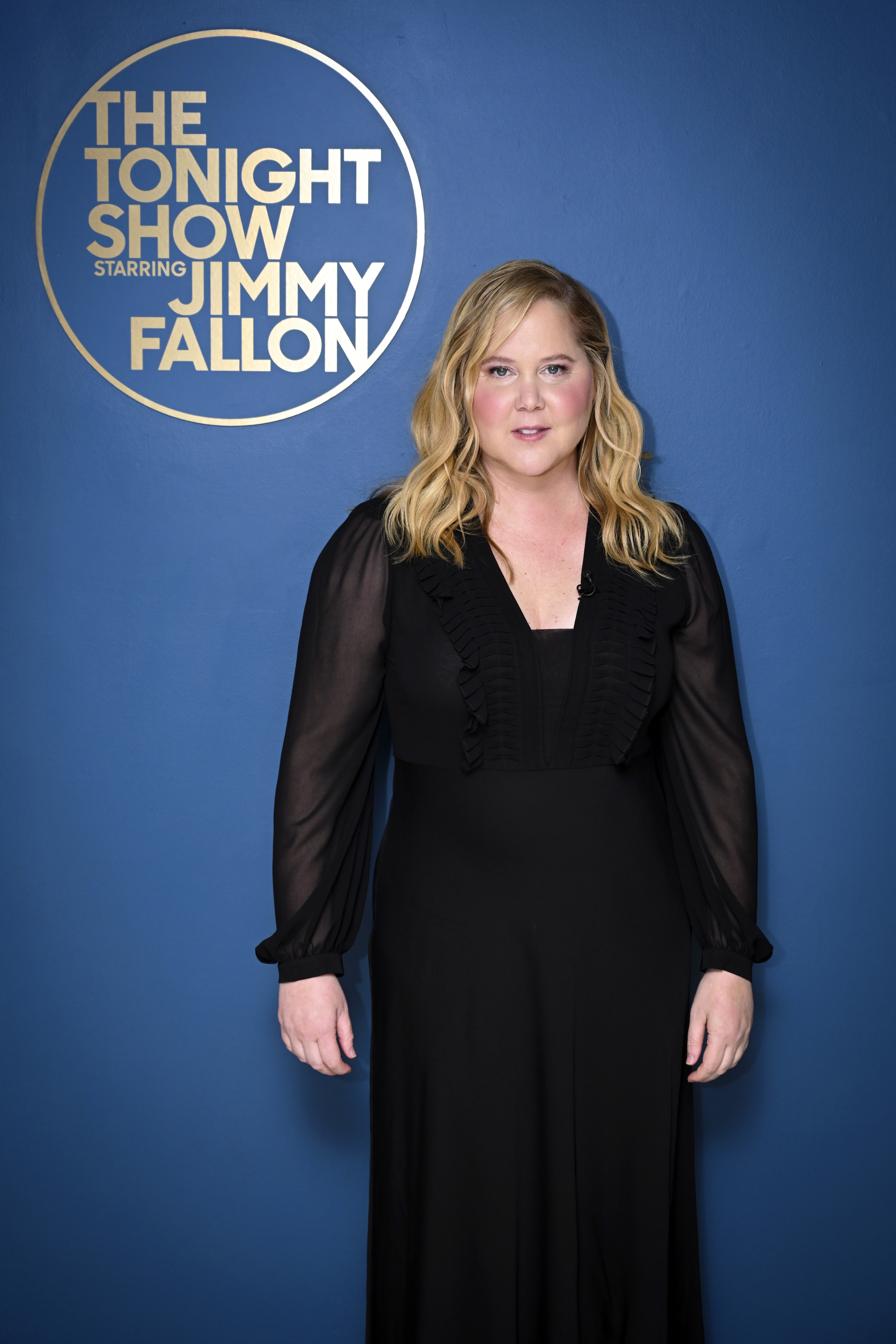 Amy Schumer stands in front of &#x27;The Tonight Show&#x27; backdrop wearing a black dress with sheer sleeves