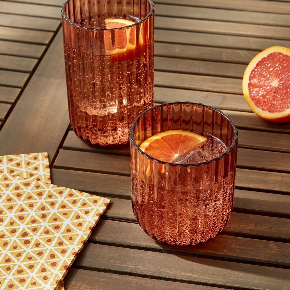 Two plastic tumblers on a wooden surface, one full with a drink and a slice of orange, the other empty, beside a cut orange