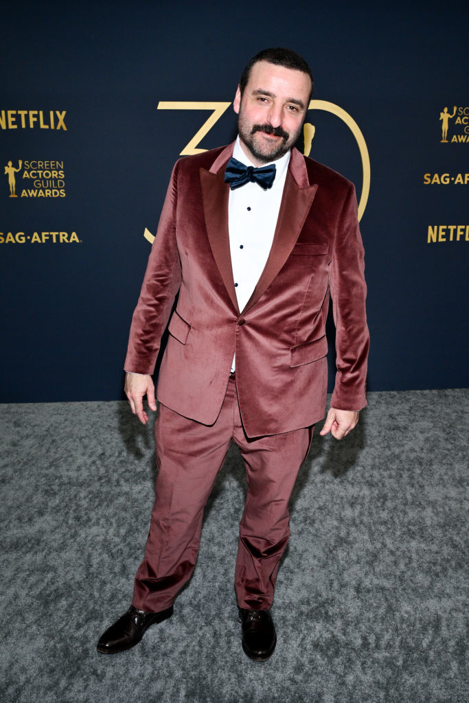 David Krumholtz wearing a maroon tuxedo, bow tie, and dress shoes