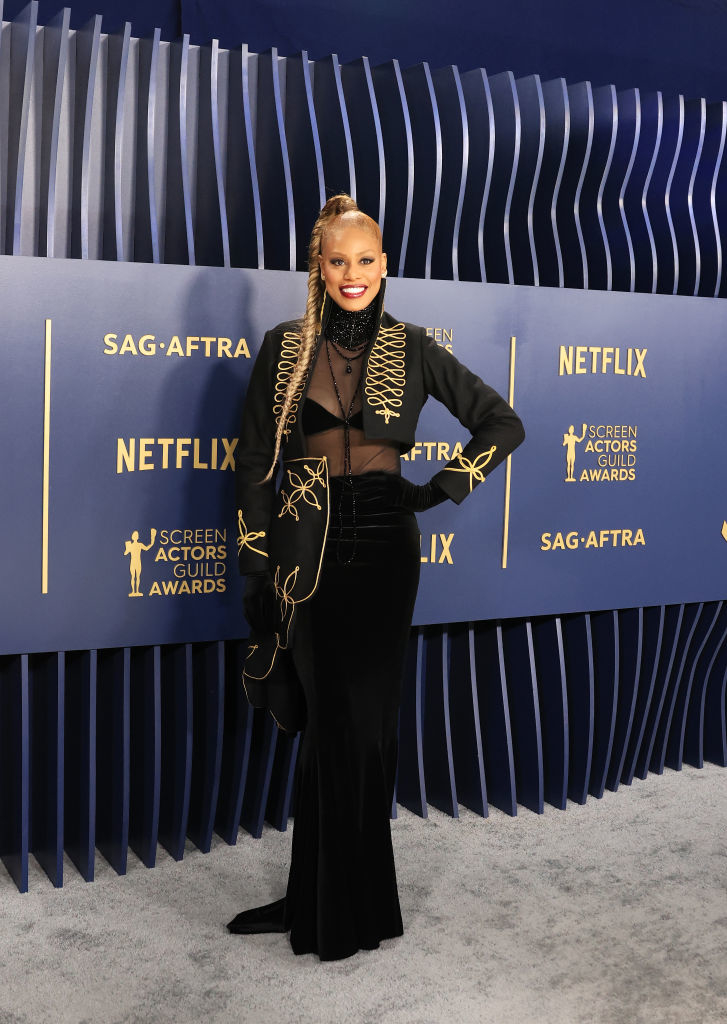 Laverne Cox stands smiling in a cropped jacket with an embroidered design over a sheer top and a long velvet skirt