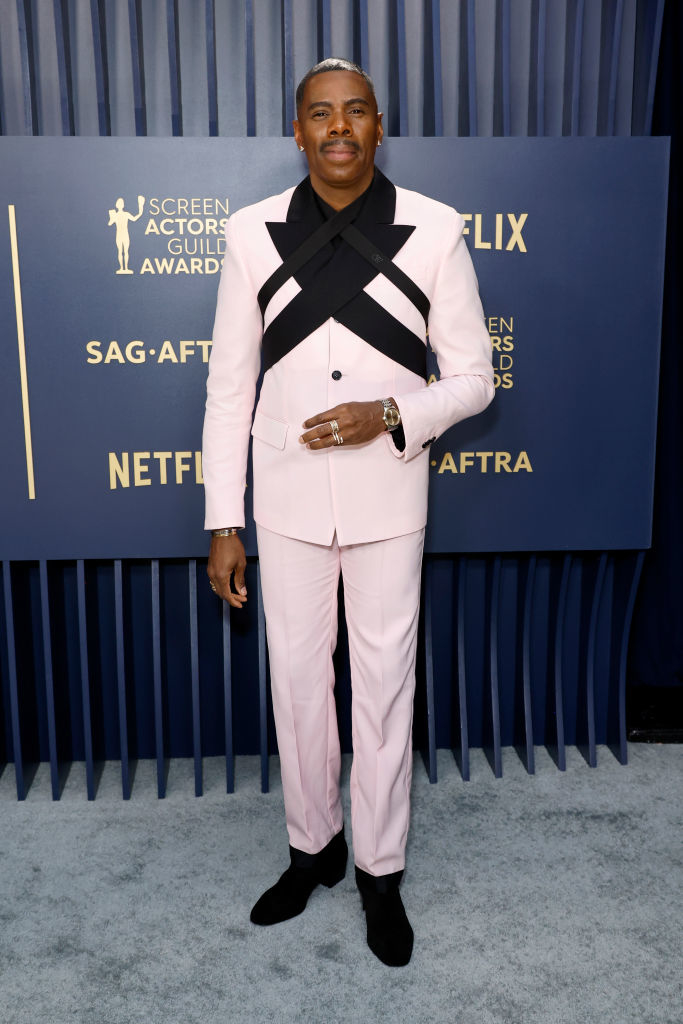 Colman Domingo on the carpet poses in a light colored suit with a unique black cross-body sash, black boots, and a watch