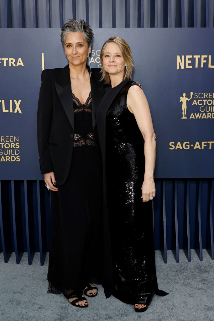 Alexandra Hedison and Jodie Foster in formal wear posing; one in a black suit and lacy top, the other in a sequined gown