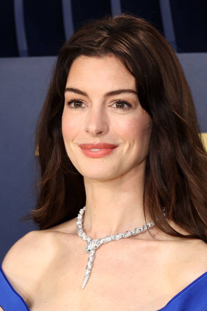 Anne Hathaway wears a blue gown with a shimmering neckpiece, smiling at an event