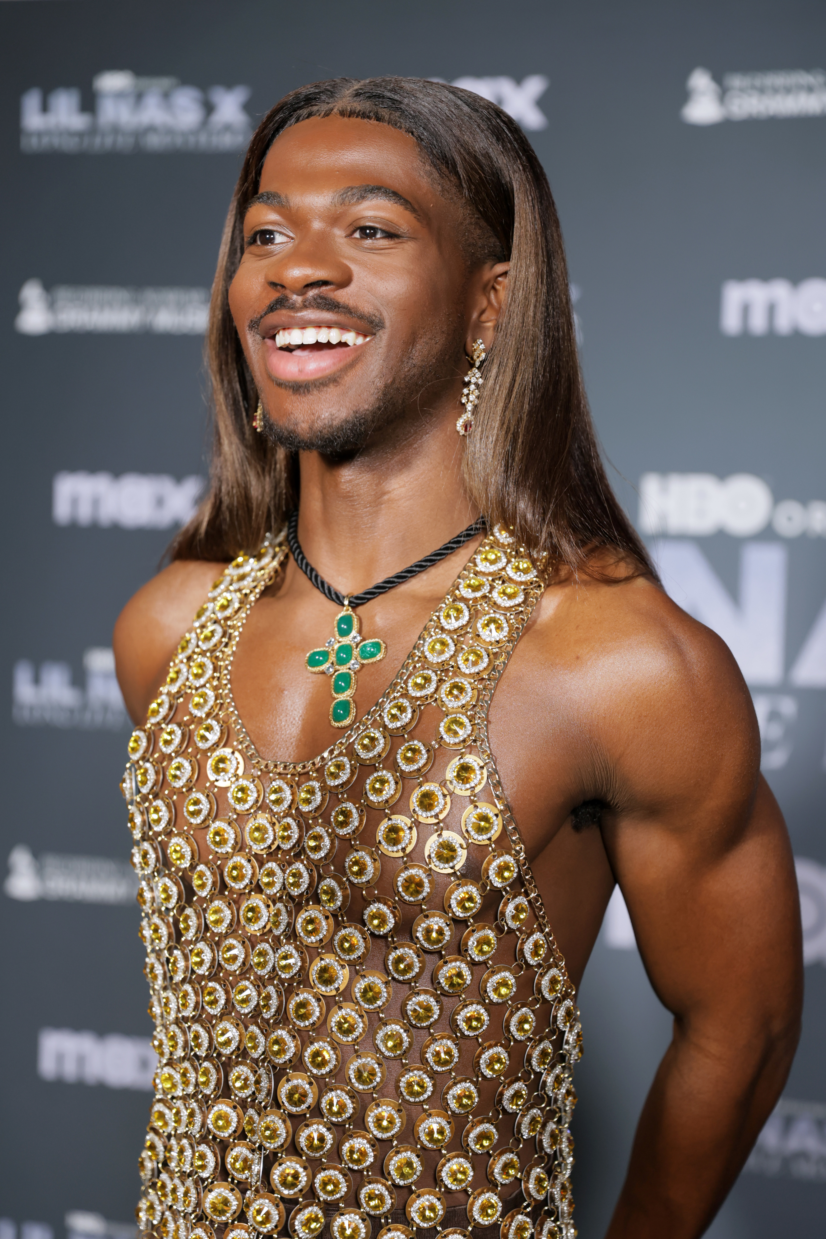 Lil Nas X smiling in an embellished sleeveless outfit with a plunging neckline at an event
