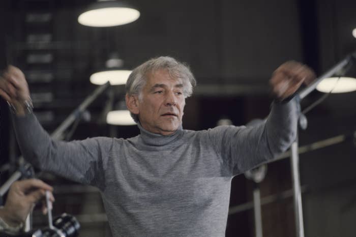 Leonard Bernstein conducting with arms raised, in casual attire, with filming equipment in the foreground
