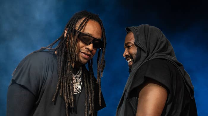 Two musicians share a joyful moment on stage; one wears sunglasses and braids, the other a hooded top