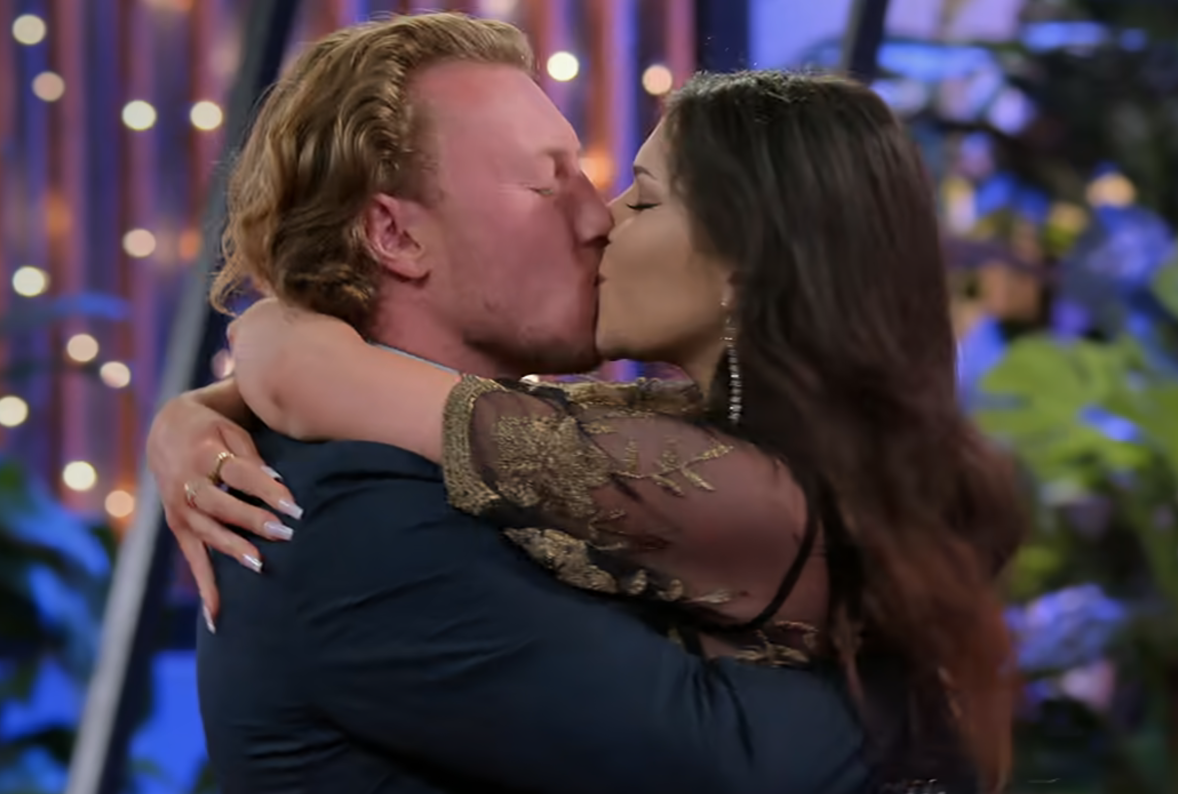 Two people embracing and sharing a kiss, surrounded by lights. The person on the left has curly hair and the person on the right has long hair