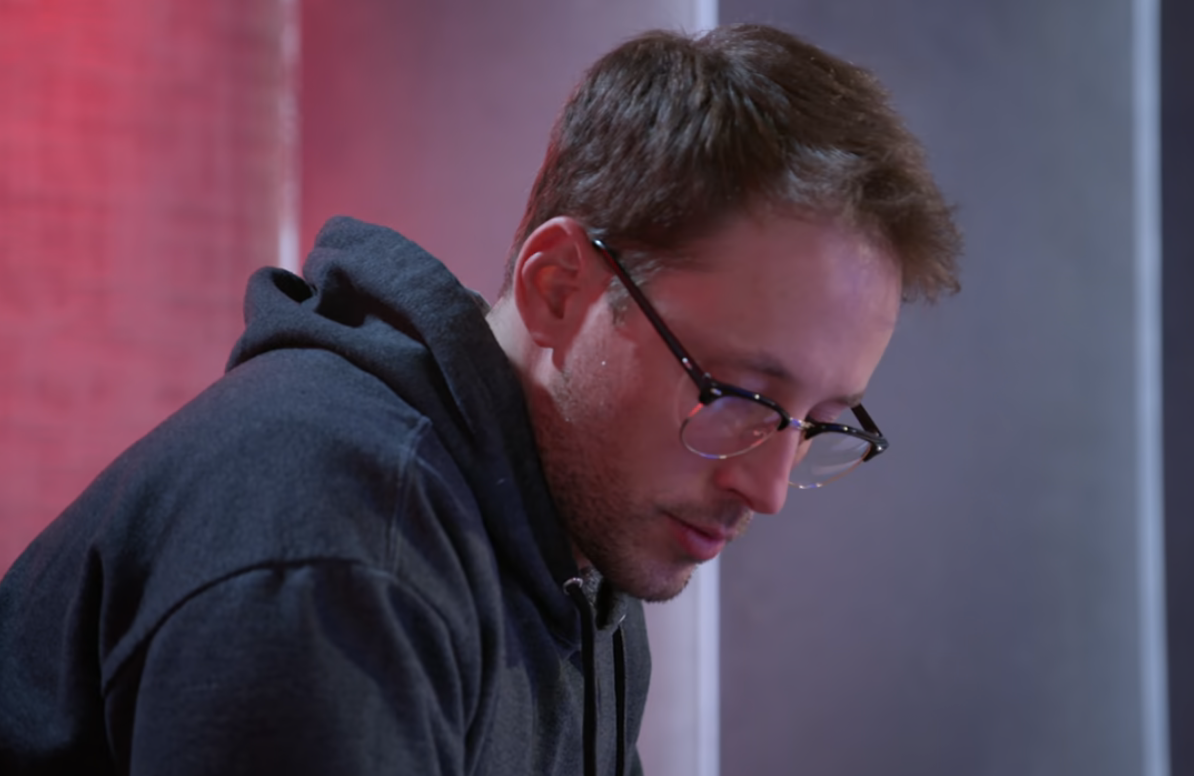 Man in glasses looking down, wearing a hoodie; indoors with a purple background