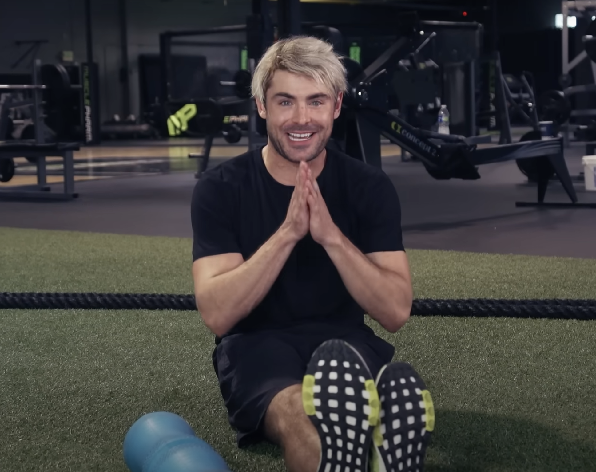 Zac smiling, sitting on gym floor with hands together, exercise equipment in background