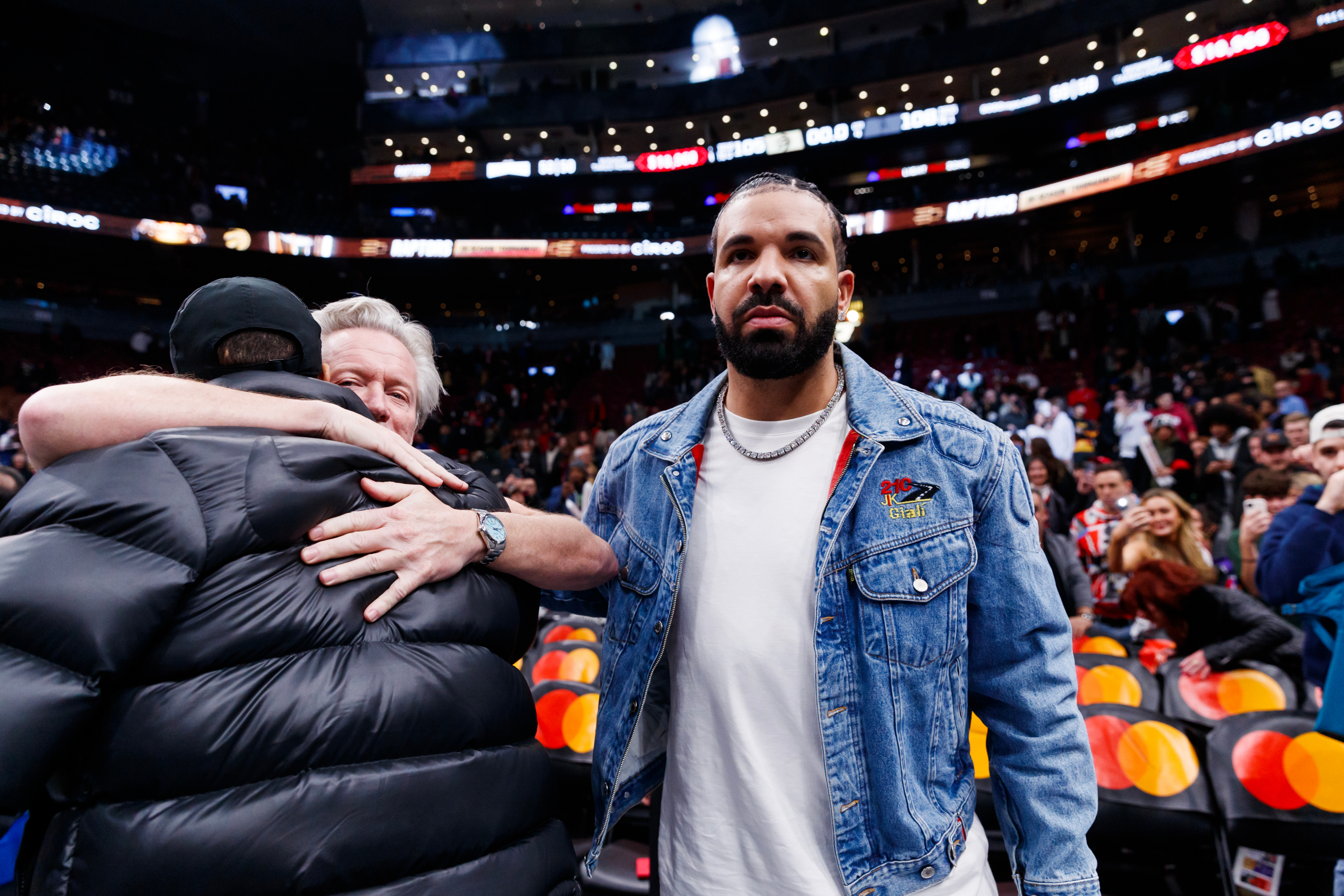 Man in a black puffer jacket hugging someone at a sports event; Drake stands by, wearing a denim jacket