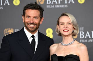 Two celebrities smiling at the BAFTA Film Awards; the person on the left in a classic suit, the right in a black off-shoulder gown with a pearl necklace