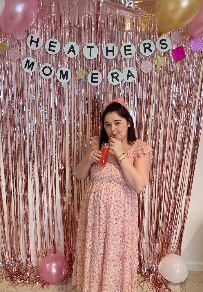 Woman in a floral dress standing in front of a pink foil backdrop with &quot;HEATHERS MOM ERA&quot; banner, sipping a drink