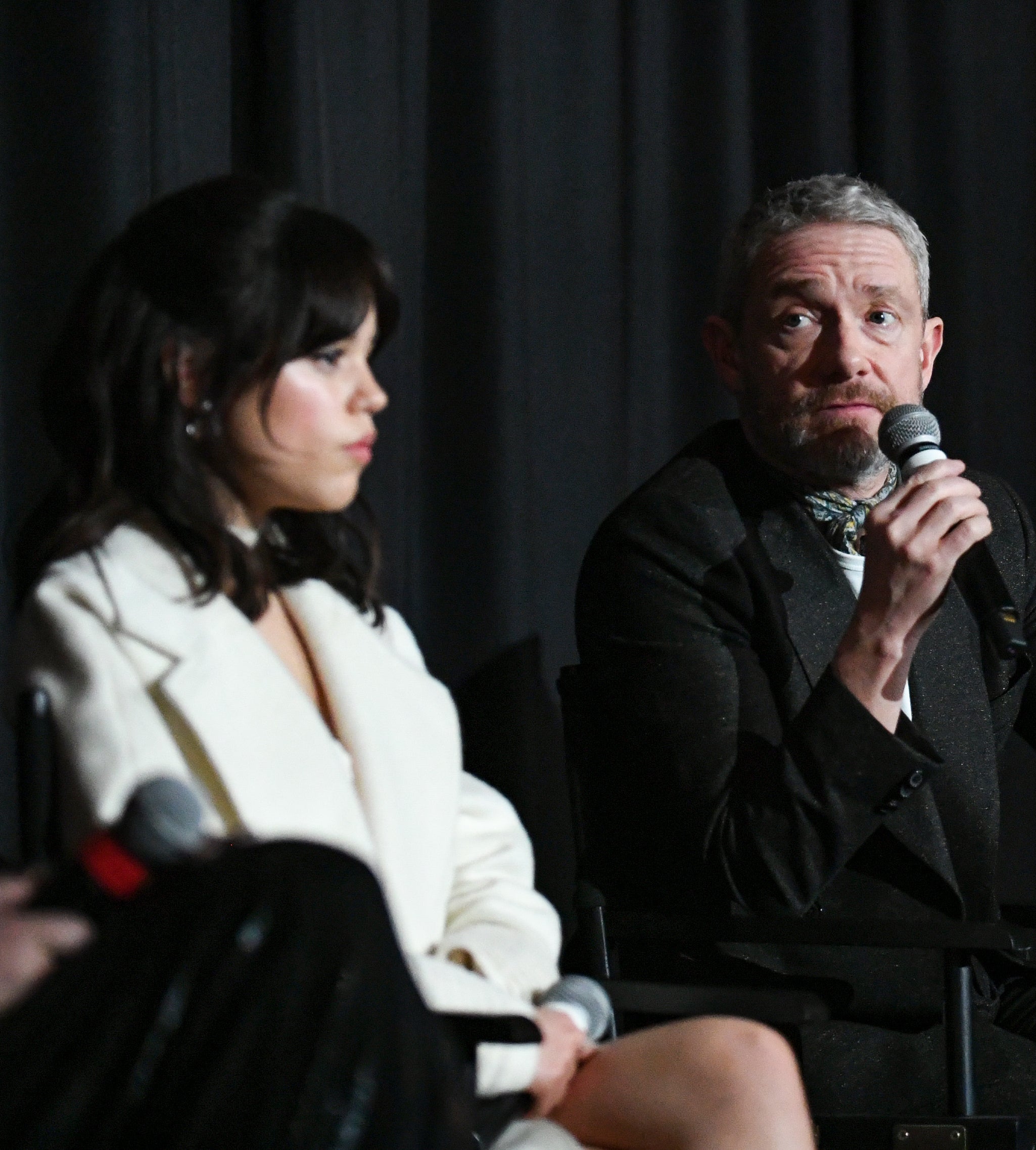 Closeup of Jenna Ortega and Martin Freeman sitting on stage as Martin speaks into a mic