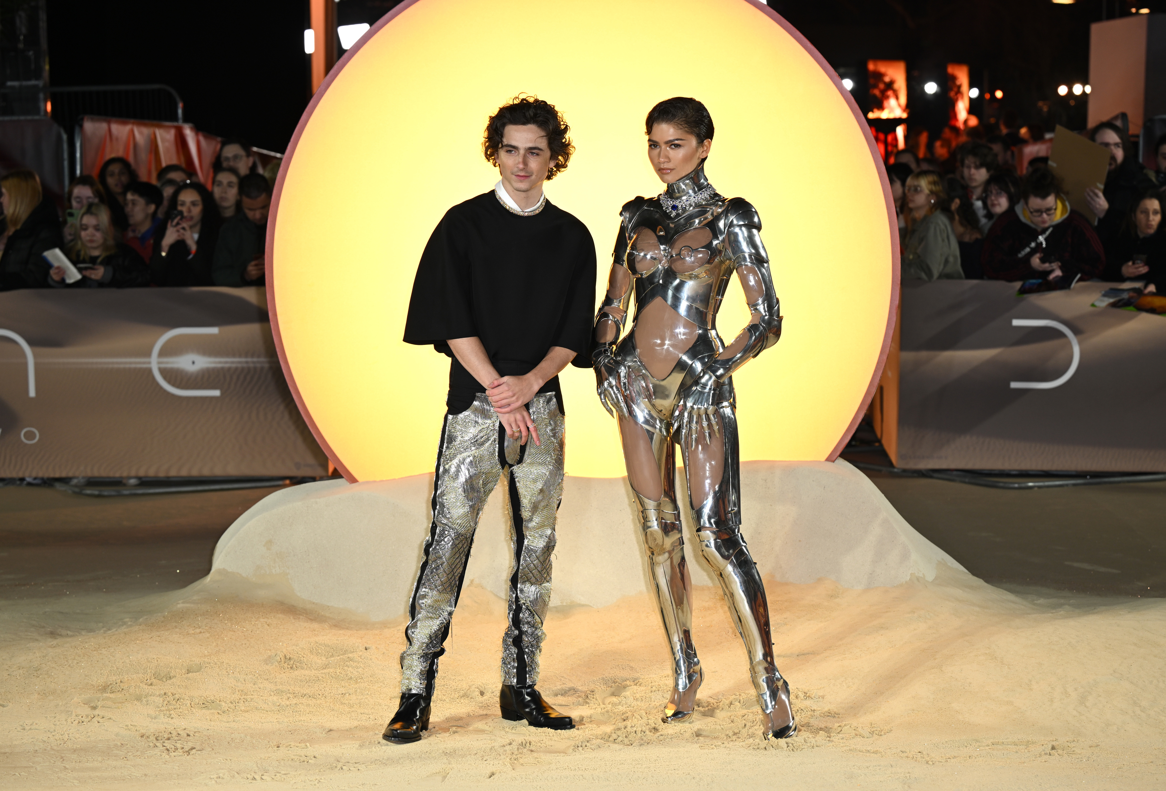 Timothée Chalamet and Zendaya on a staged area, he in a black top with metallic pants, and she in a glossy sculptural outfit
