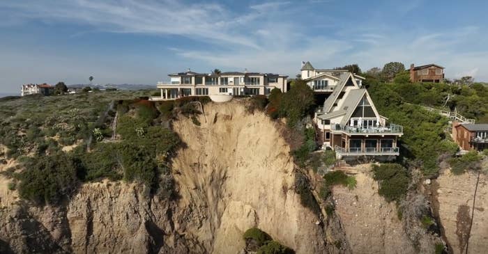 Coastal homes on cliff edge with evident erosion, under clear skies