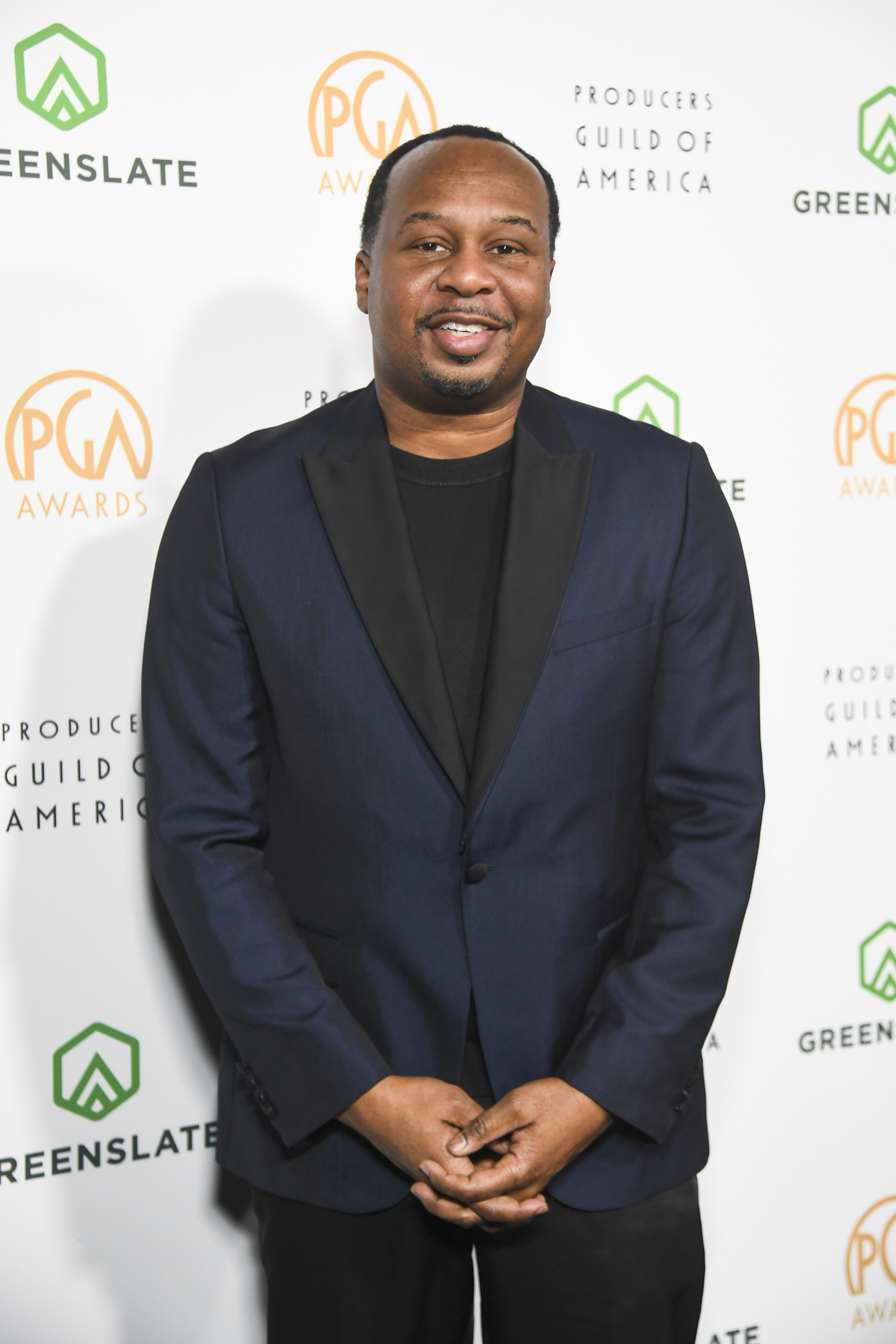 Roy smiles in a suit at the Producers Guild Awards backdrop