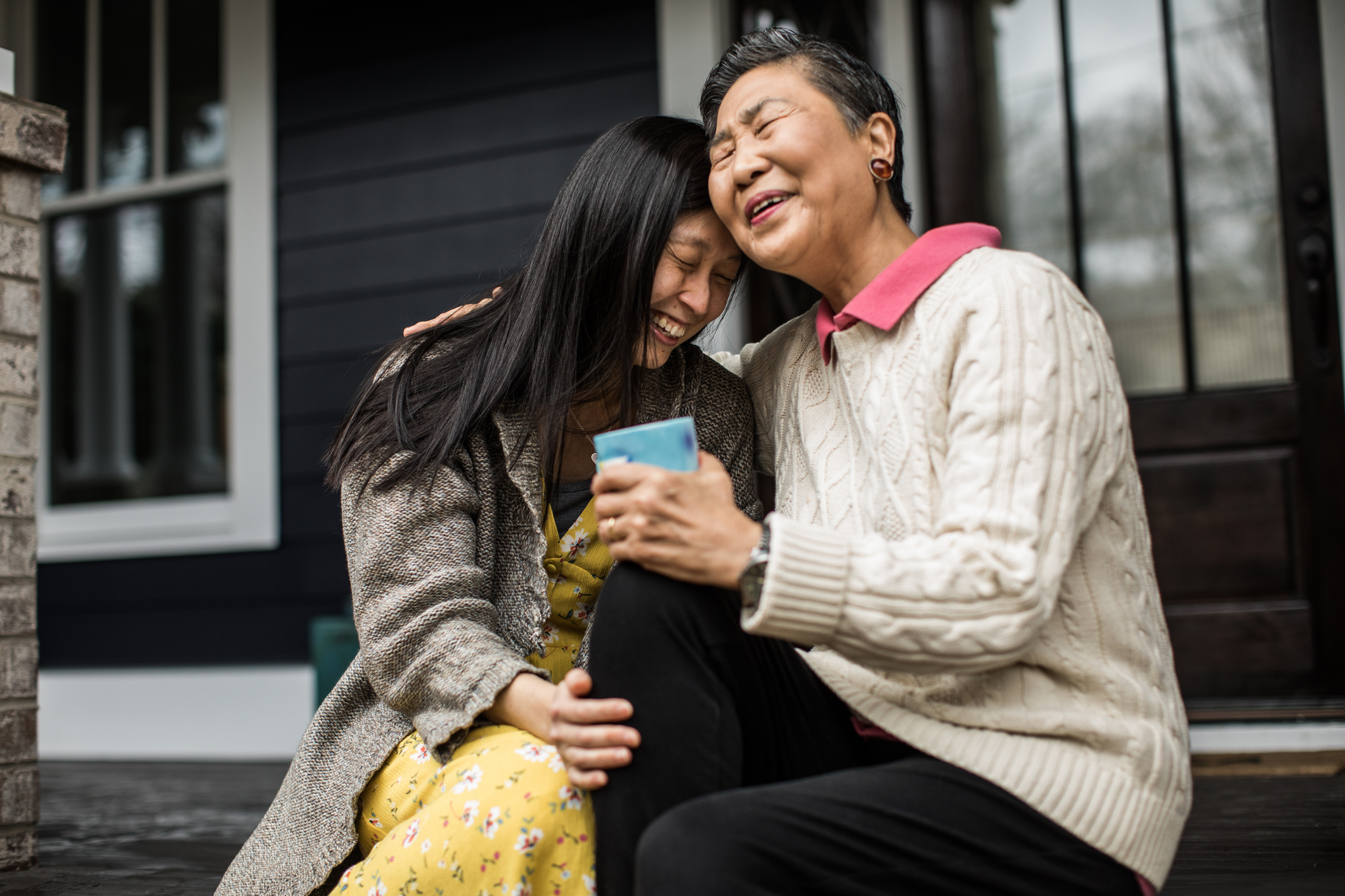 Two people smiling and embracing while sitting on a porch step, one holding a smartphone