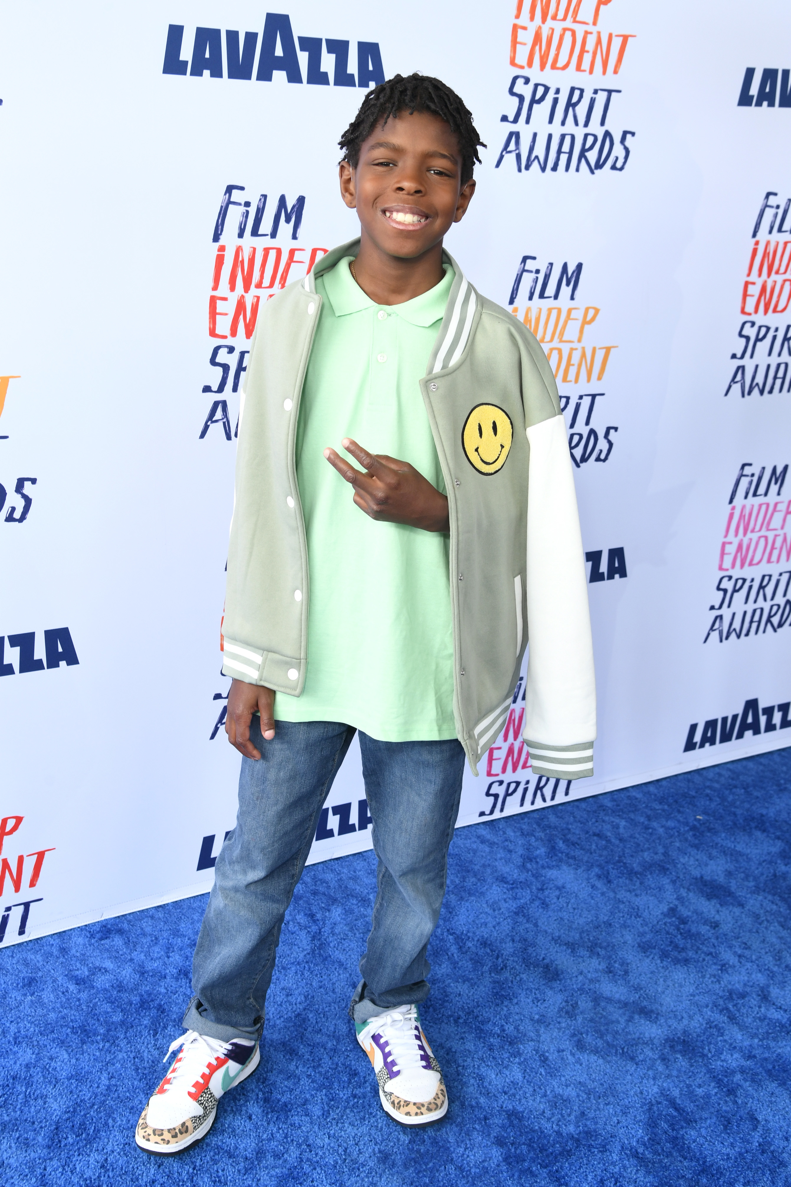 Keivonn Woodard stands on the red carpet with a casual layered look, a smiley-face detail on outerwear, and patterned sneakers