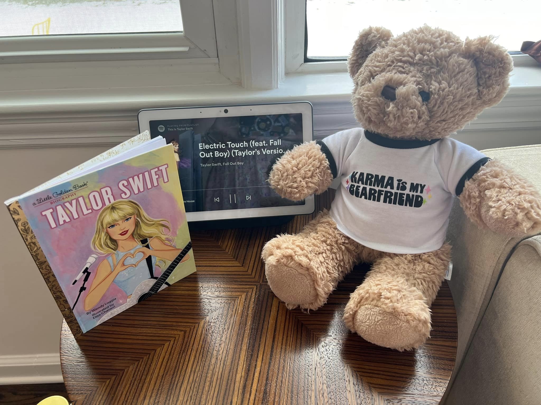 A Taylor Swift little golden book, a tablet playing “Electric Touch”, and a teddy bear in a “KARMA IS MY BOYFRIEND” shirt