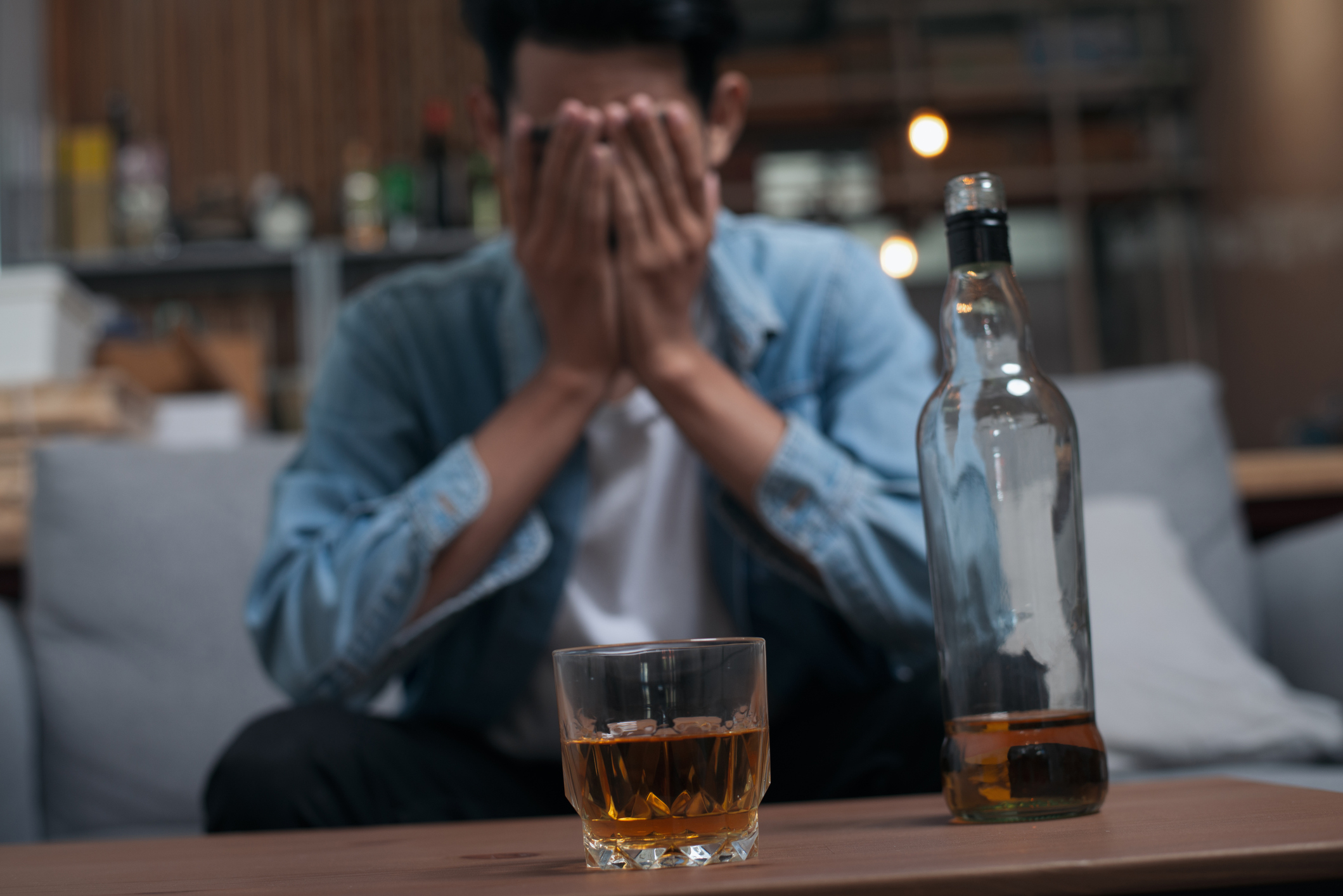 Person sitting with hands covering face, a bottle and a glass of whiskey in the foreground, signifying distress or sadness