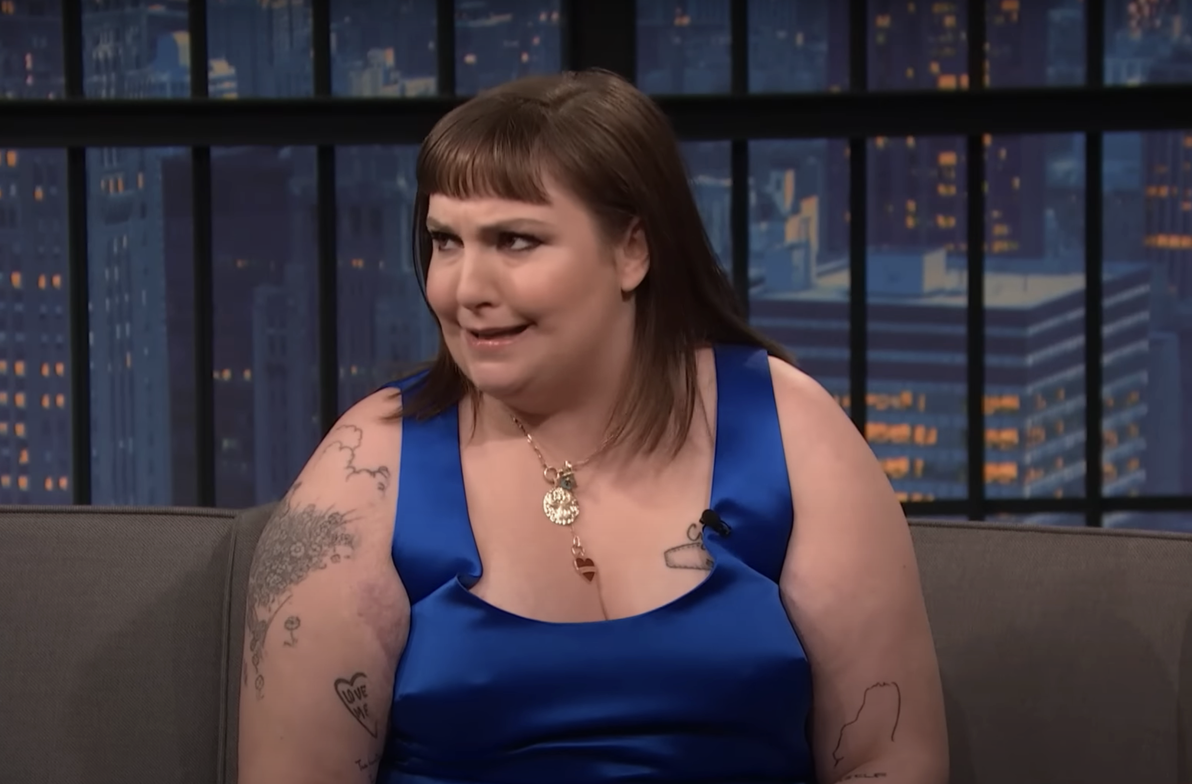 Lena in a sleeveless dress and necklace on talk show, tattoos on arms, smiling