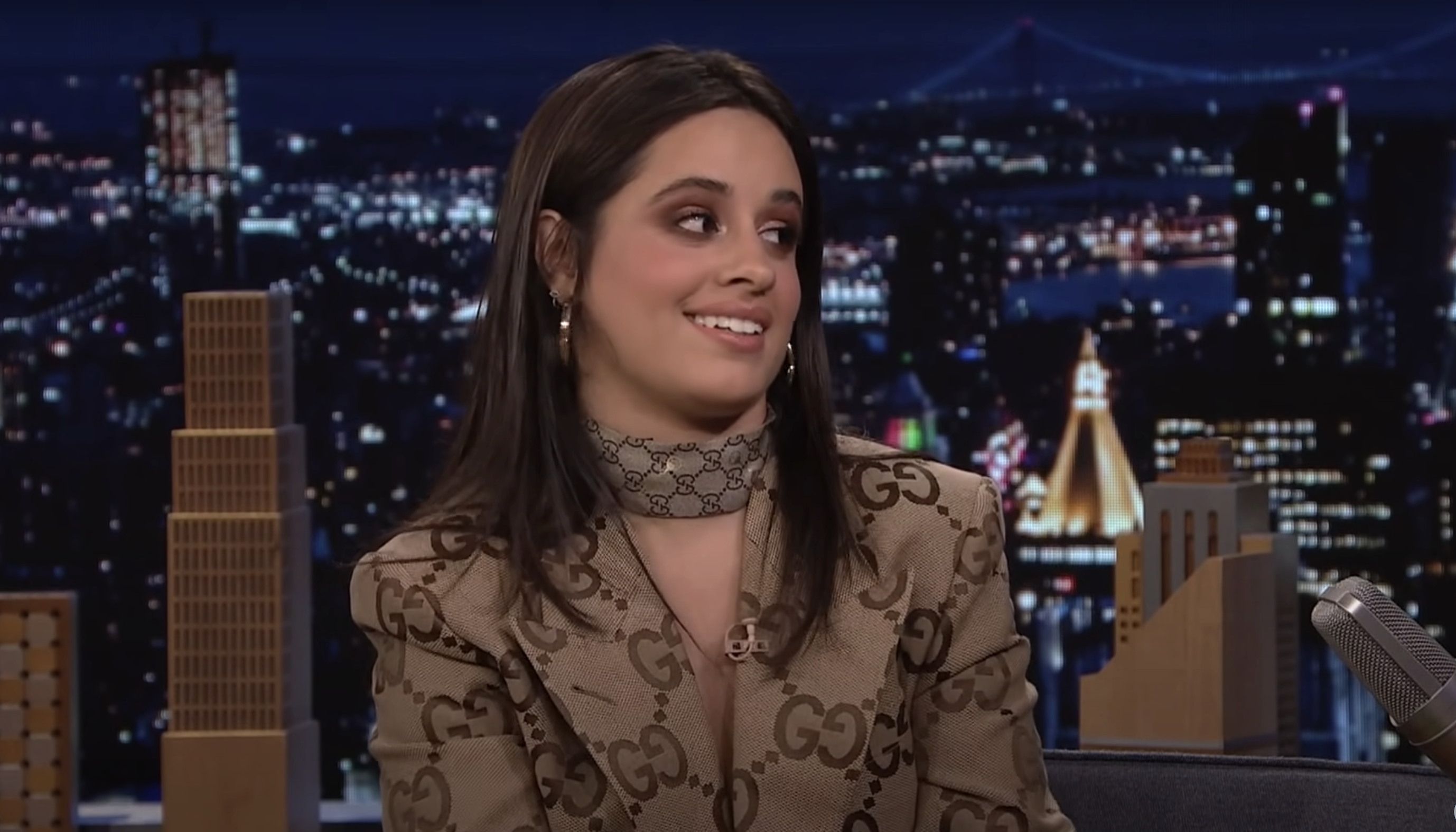 Camila smiling, in patterned outfit with choker, on a talk show set