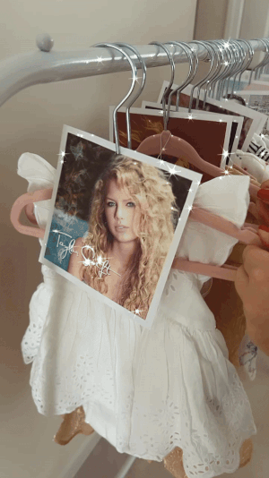 Hand holding Taylor Swift&#x27;s &quot;Fearless&quot; album cover on a hanger with white dresses in the background