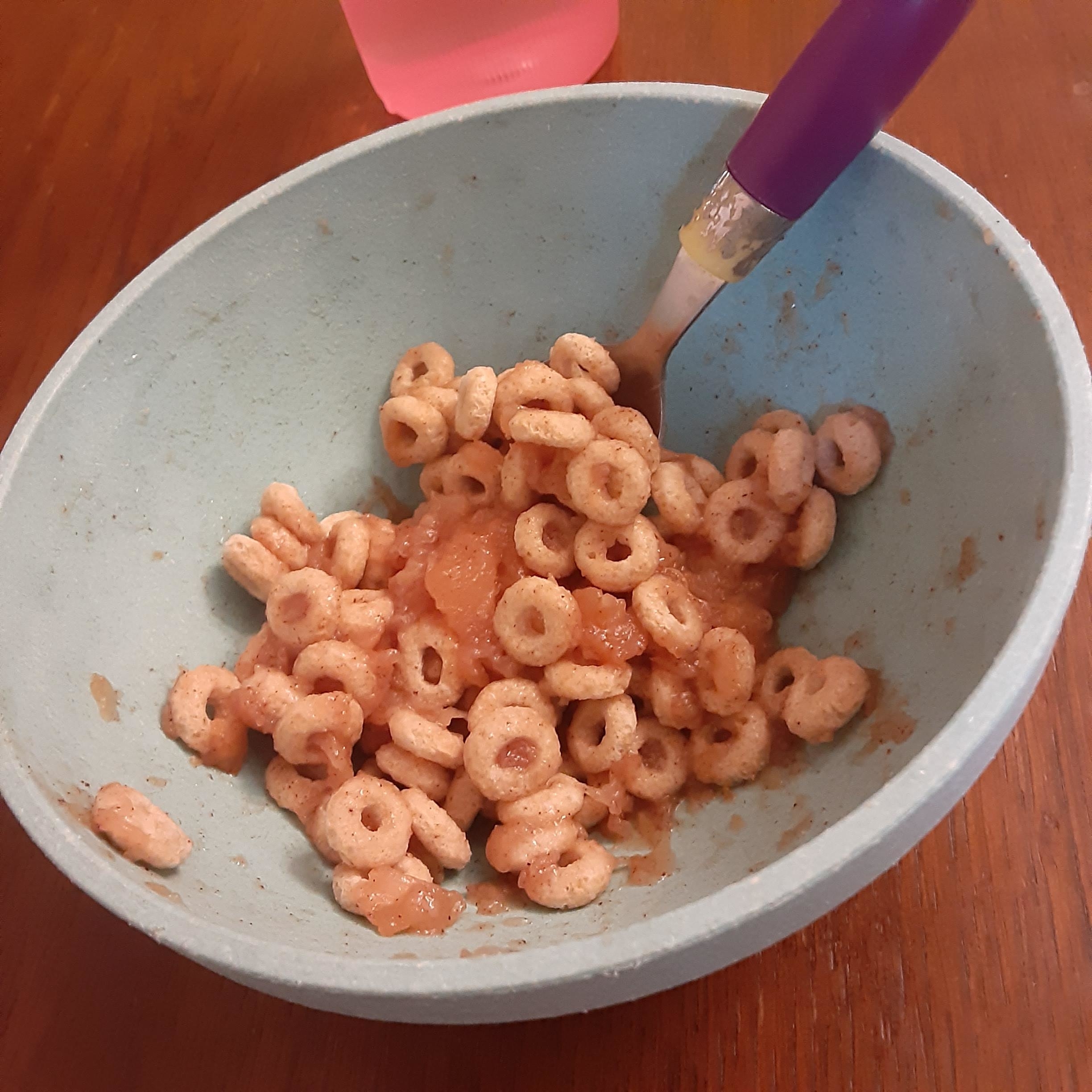 A bowl of cereal with applesauce and a spoon, partially eaten