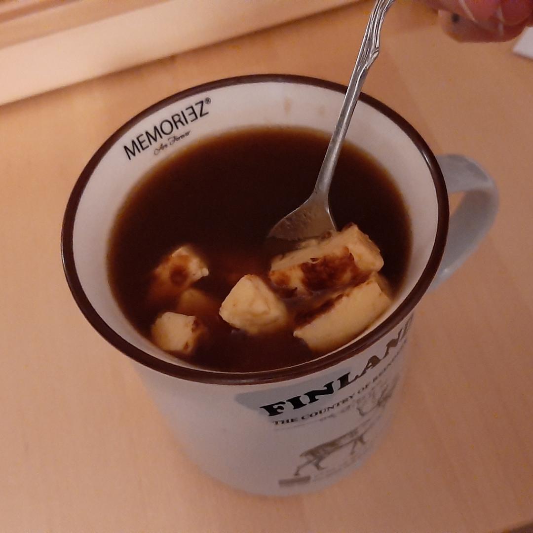 A cup of black coffee with floating chunks of cheese, being stirred with a spoon