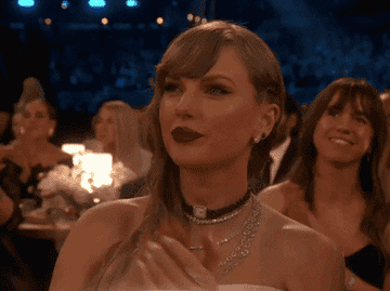 Taylor swift clapping and smiling at an event with attendees in the background