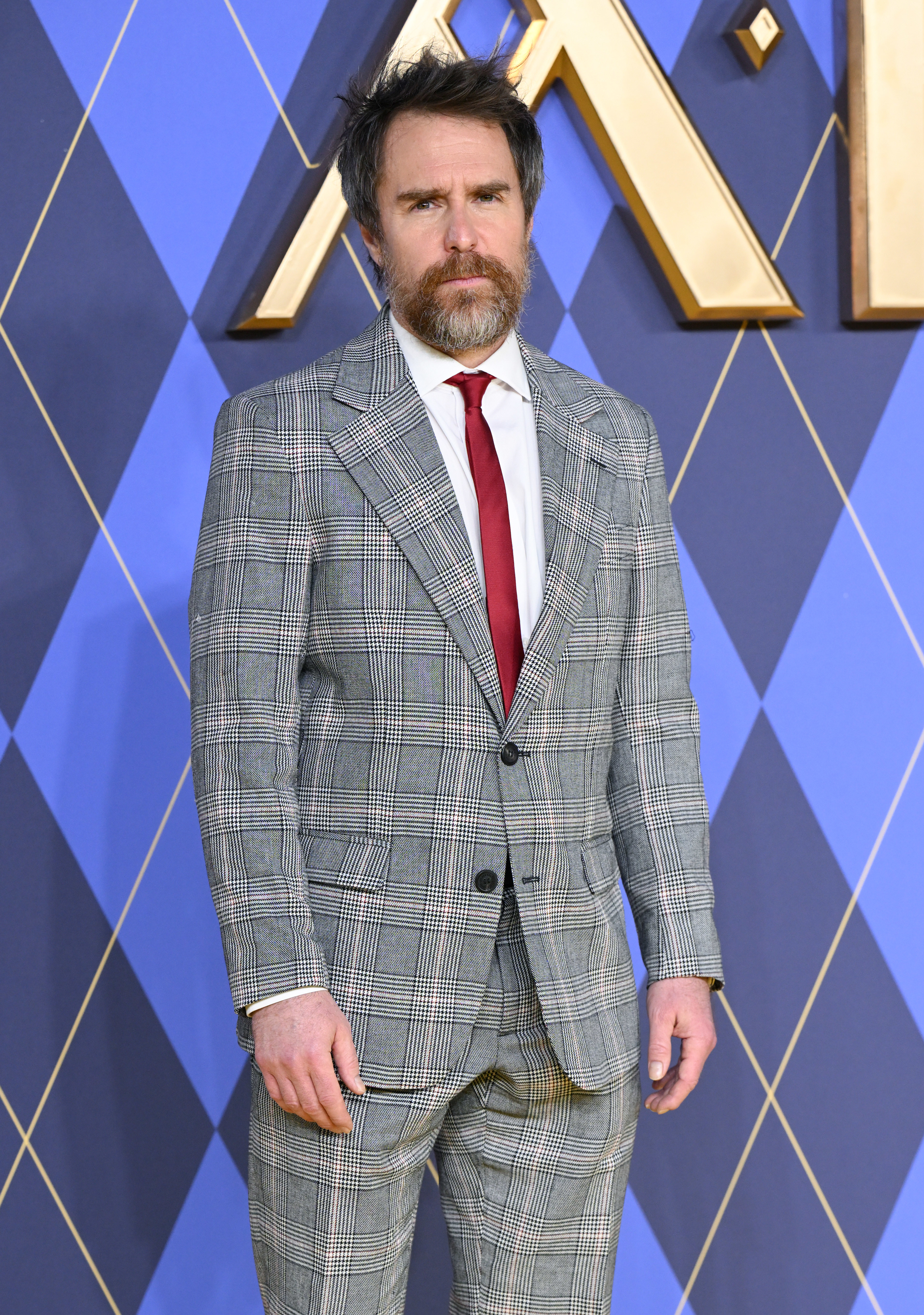 Sam Rockwell stands on the carpet in a plaid suit with a tie