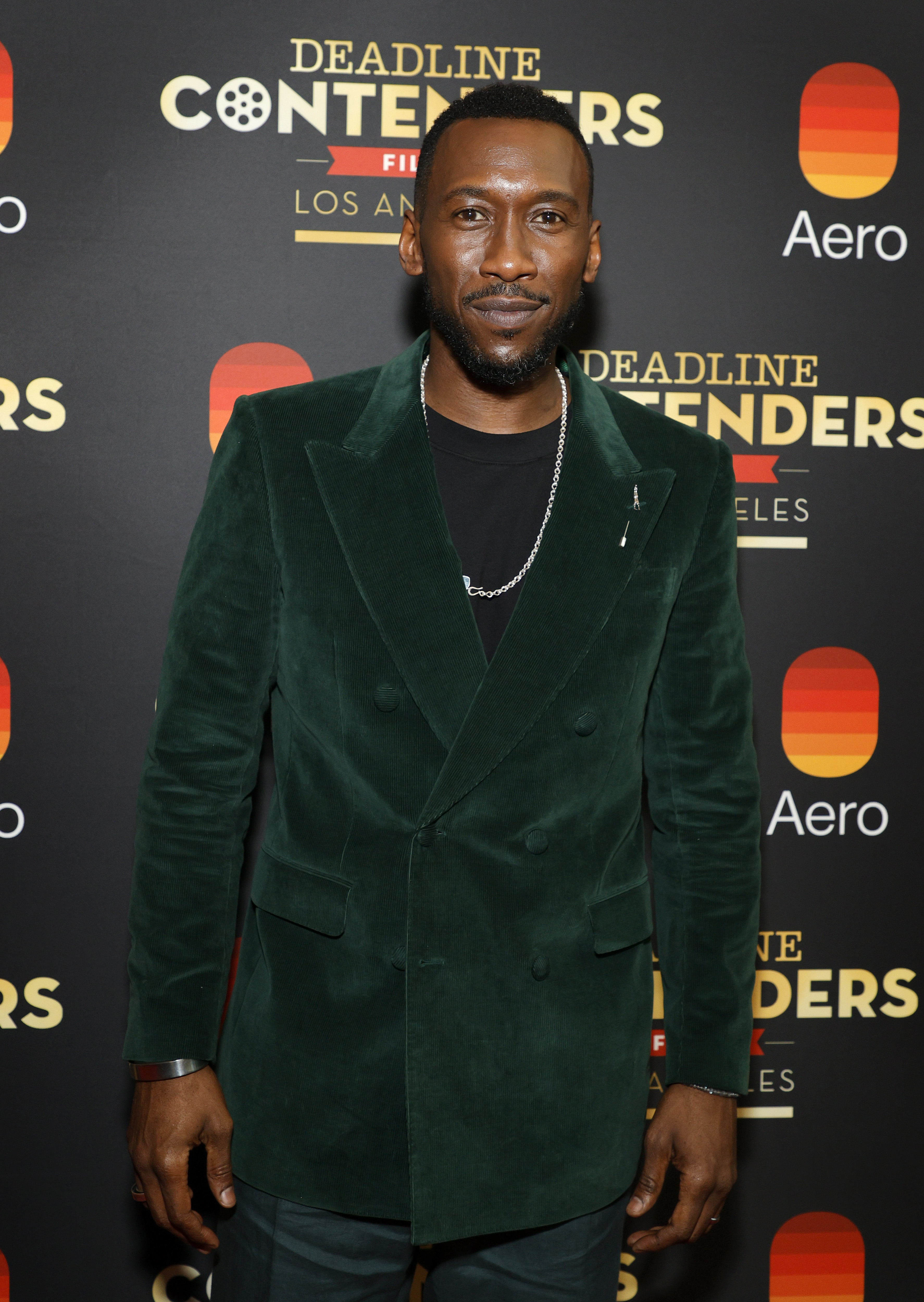 Mahershala Ali stands on red carpet event wearing formal velvet suit with lapel pin