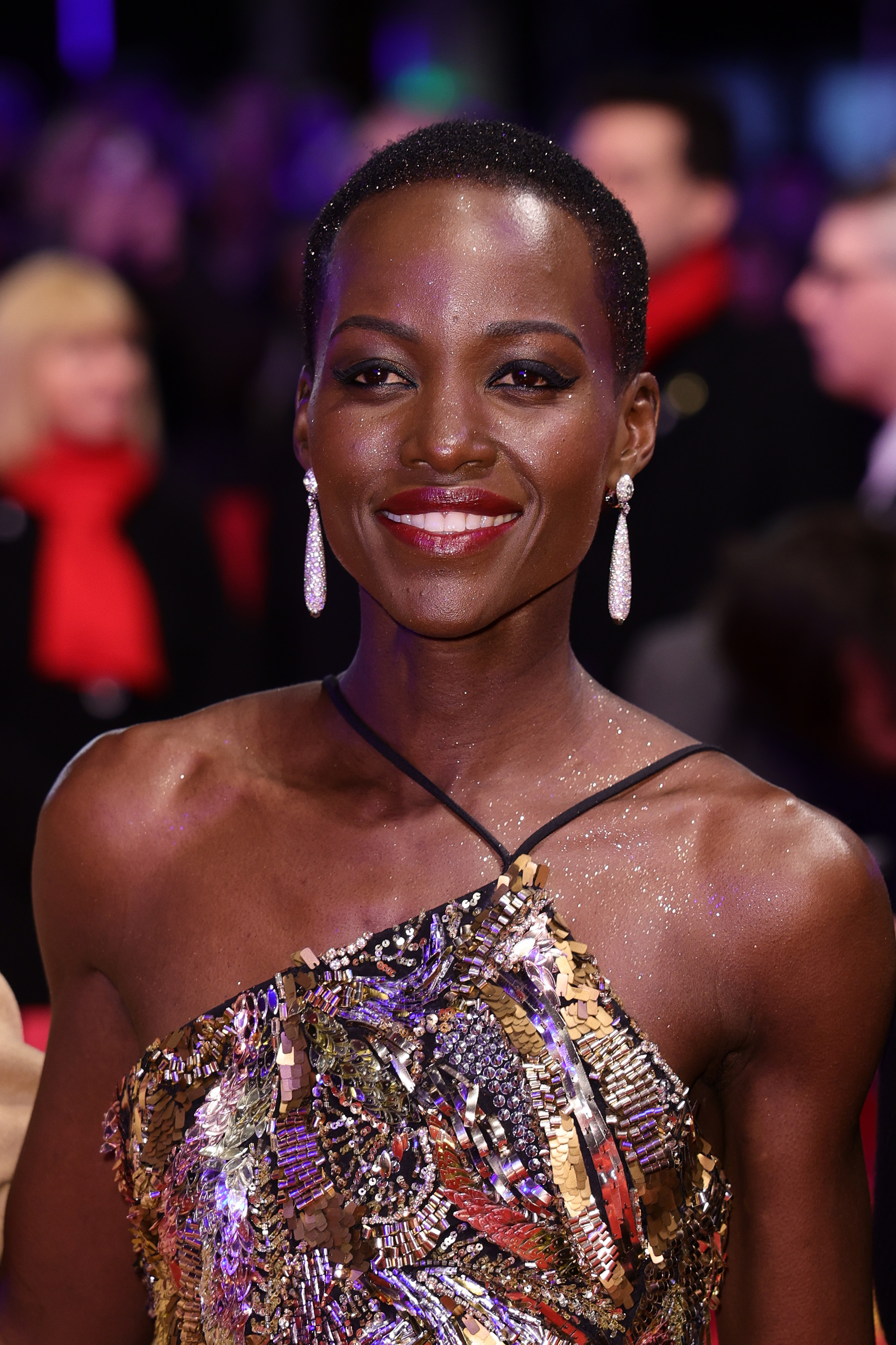 Lupita Nyong&#x27;o smiling in a metallic dress with sequins and long, dangling earrings on the red carpet