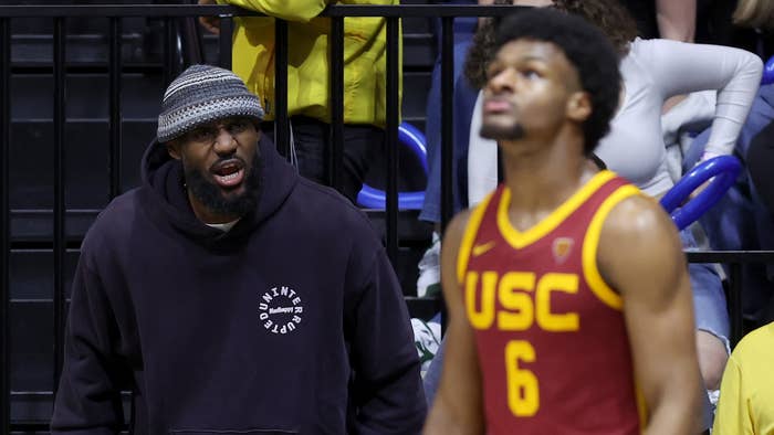 Two individuals at a basketball game, one in a hoodie and beanie speaking, the other in a USC basketball uniform listening
