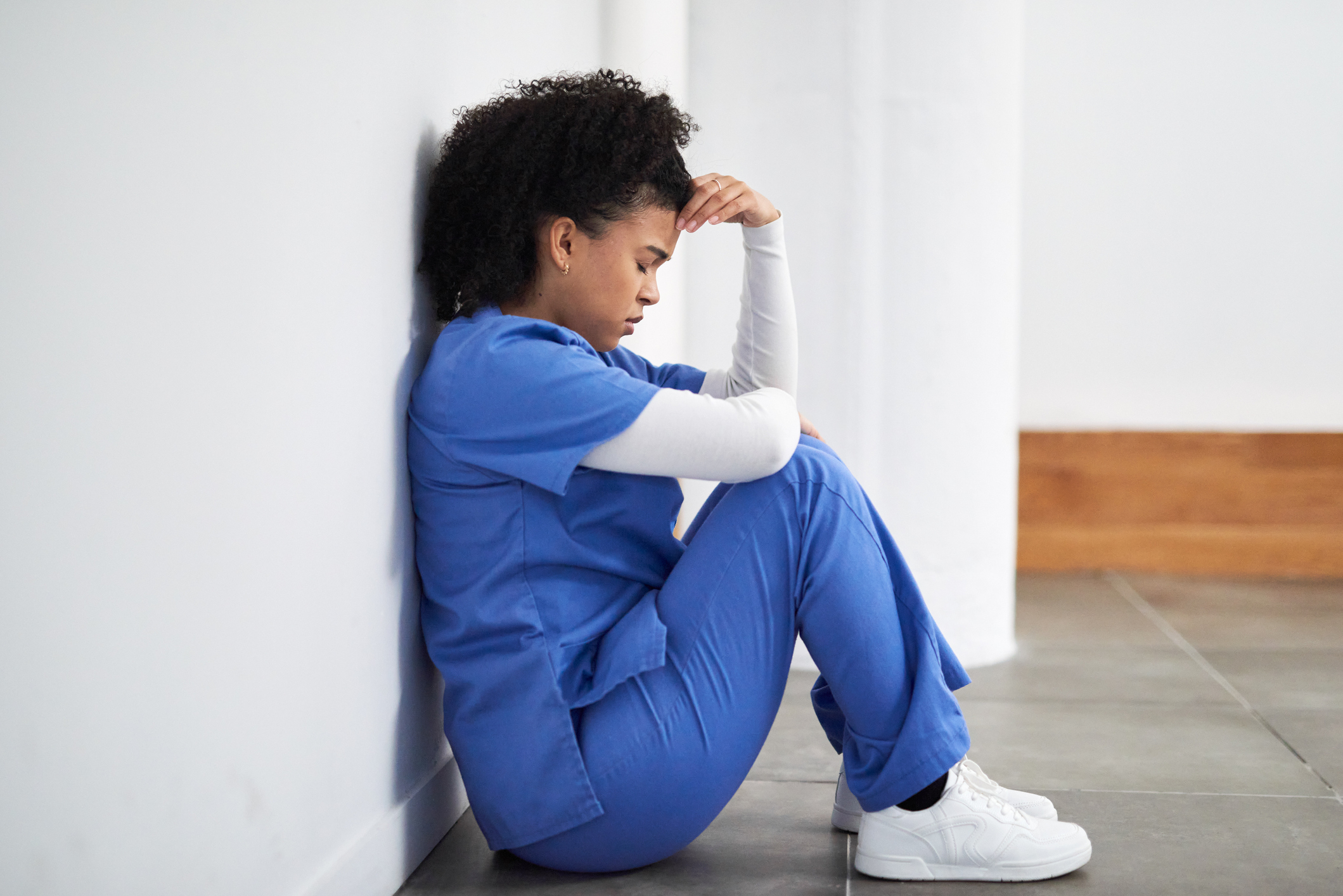 A person in medical scrubs sits on the floor, leaning against a wall in a pensive pose