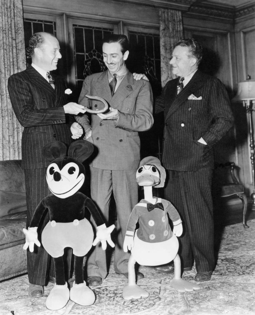 Walt Disney with Mickey Mouse and Donald Duck cutouts, showing an award to two men in suits
