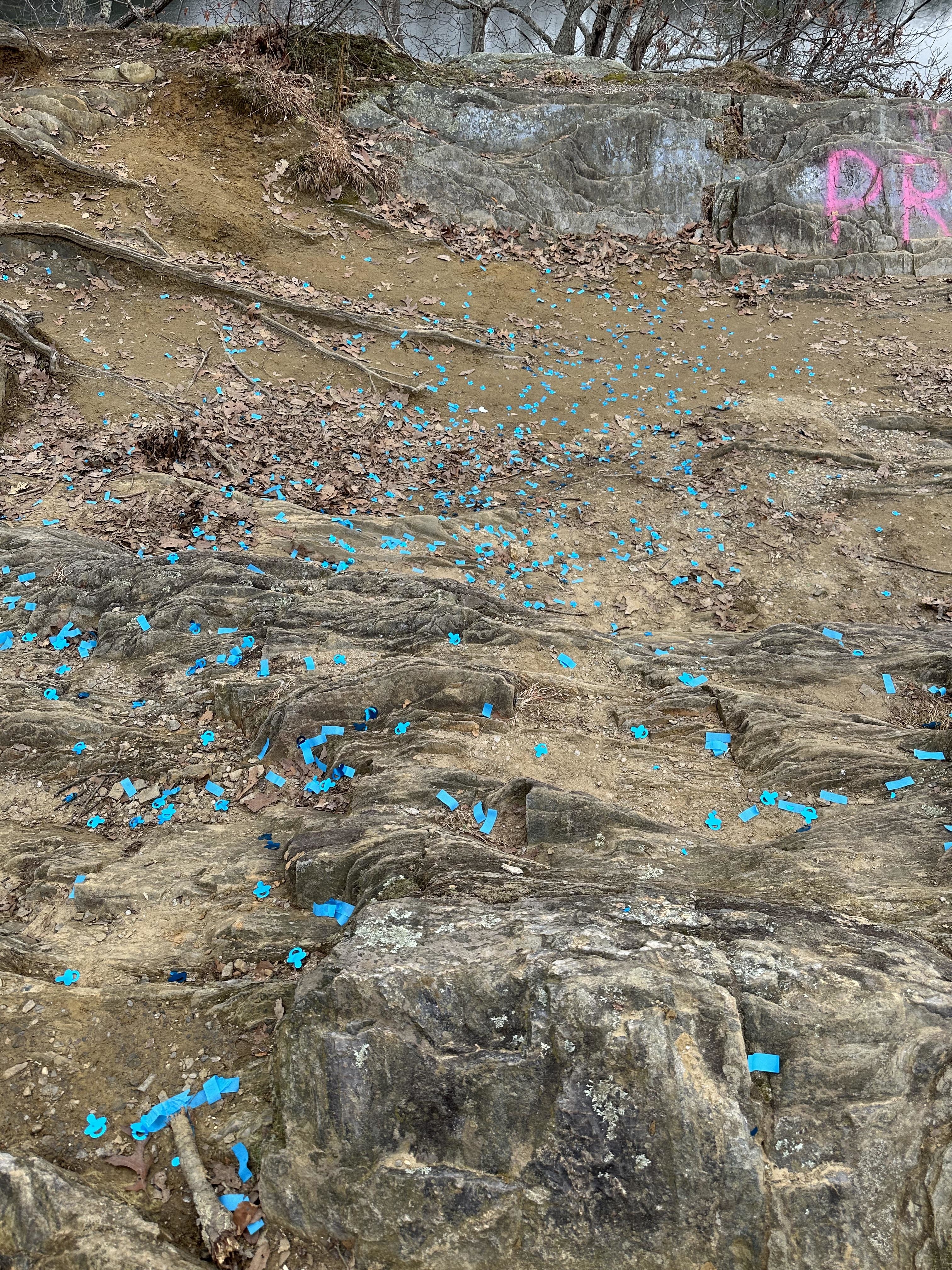 Scattered pieces of blue confetti on a rocky ground with sparse vegetation and graffiti in the background