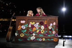 sabrina carpenter and taylor swift smile at each other while seated behind a floral painted piano on stage