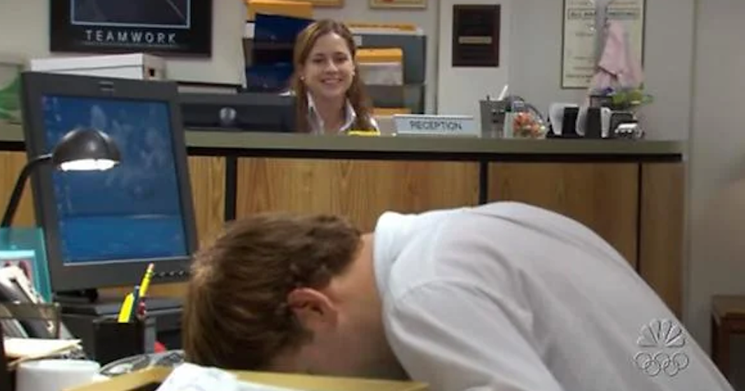 Man sleeping at desk with amused woman looking on, in an office setting from a TV show scene