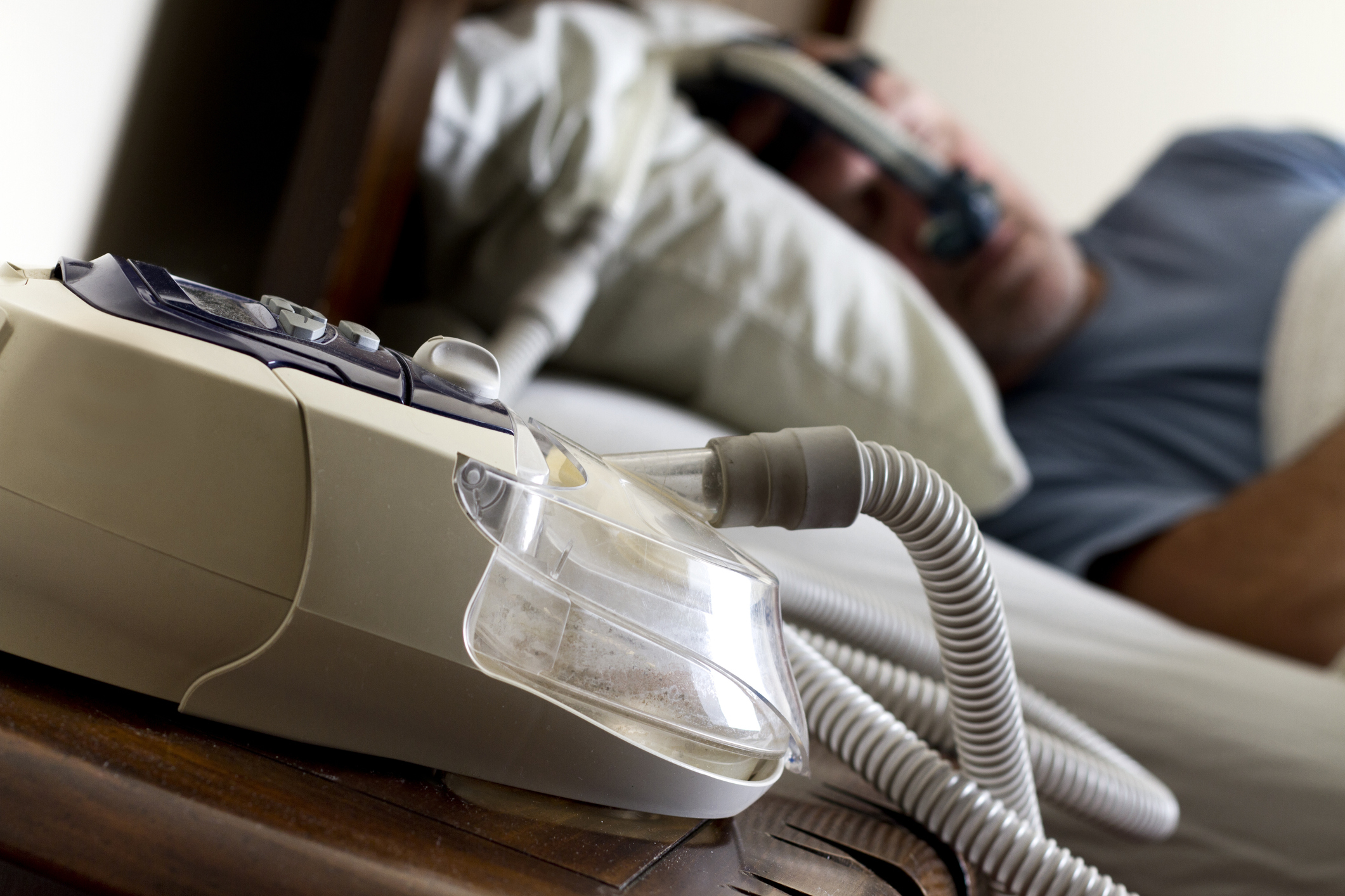 A CPAP machine on a nightstand with a person lying in bed in the background wearing the mask