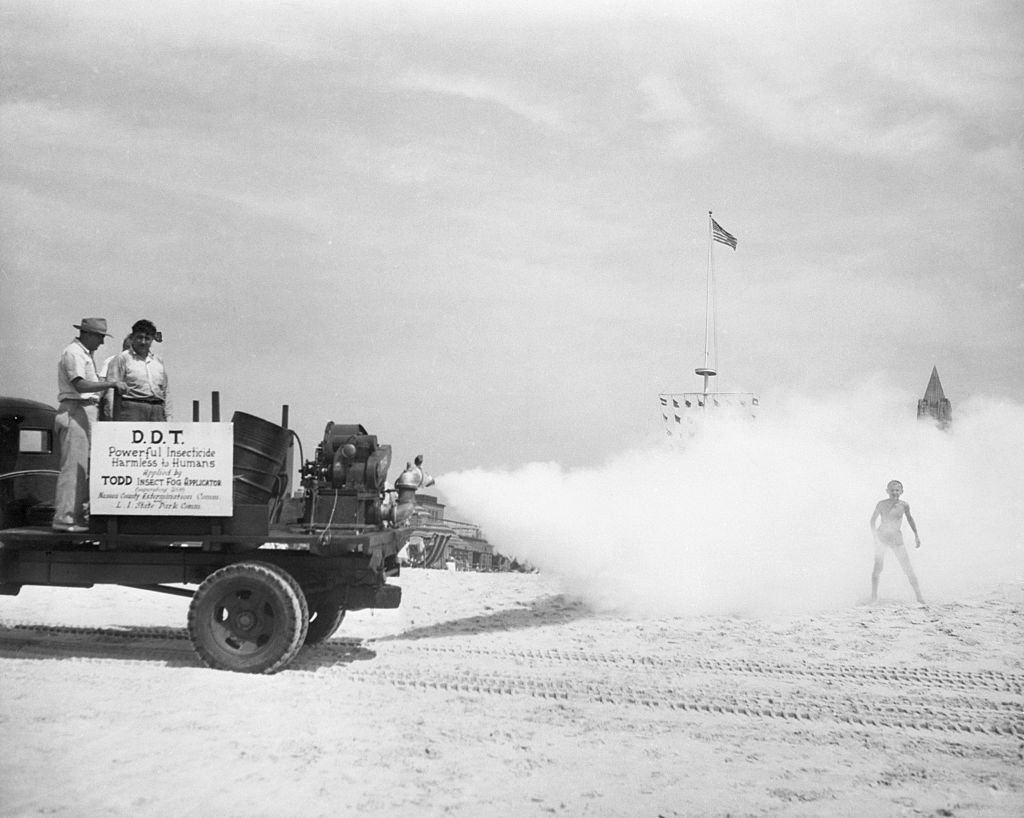 Vintage photo of a truck spraying DDT on a beach to control mosquitos, with people and a ship in the background