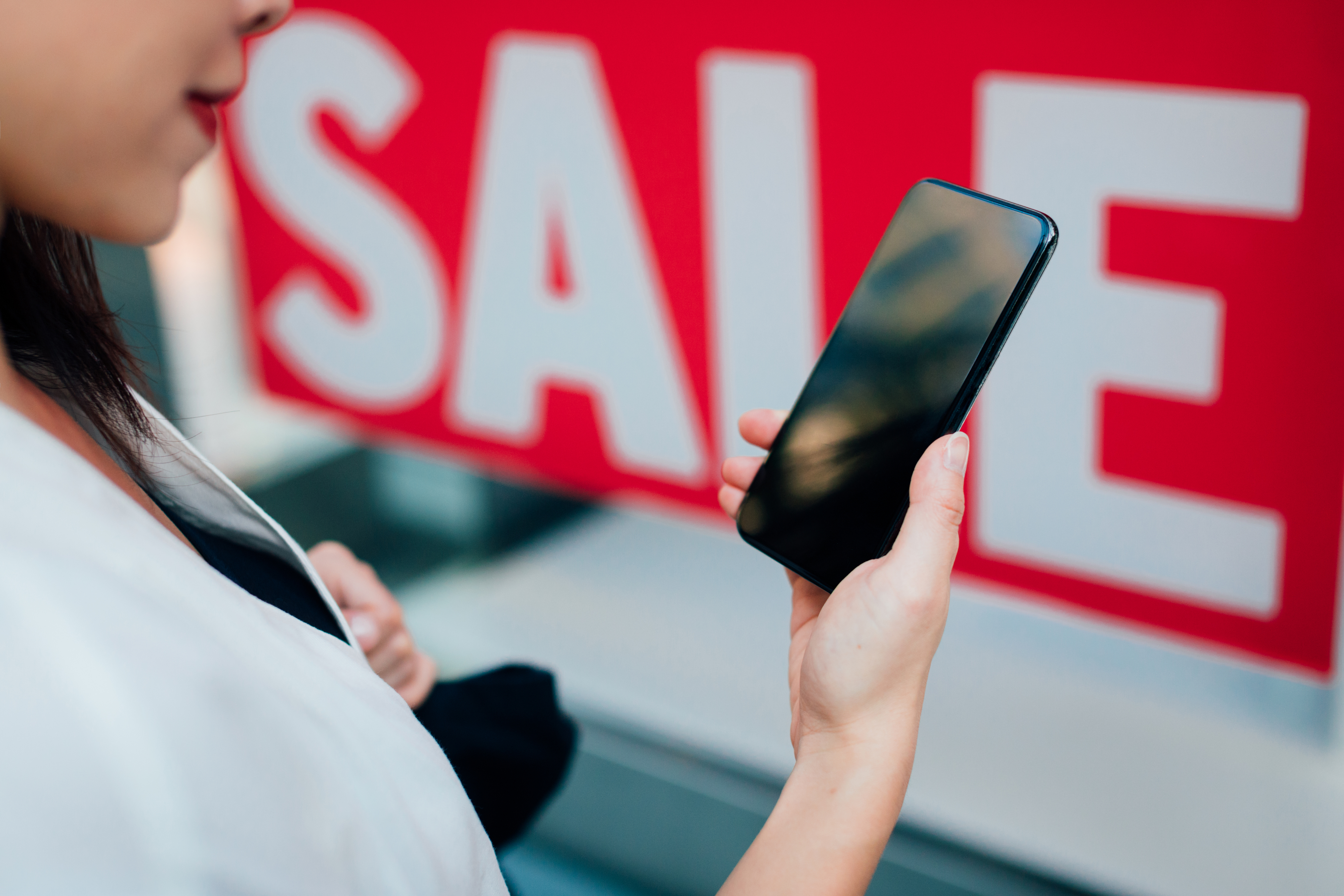 Person using smartphone, standing near a &quot;SALE&quot; sign, reflecting mobile shopping and promotions