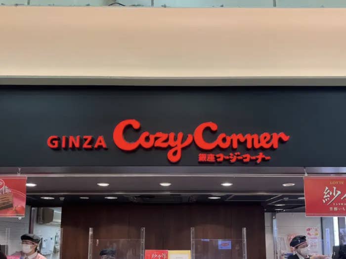 Storefront sign for &quot;GINZA Cozy Corner&quot; above entrance, with employees wearing masks visible