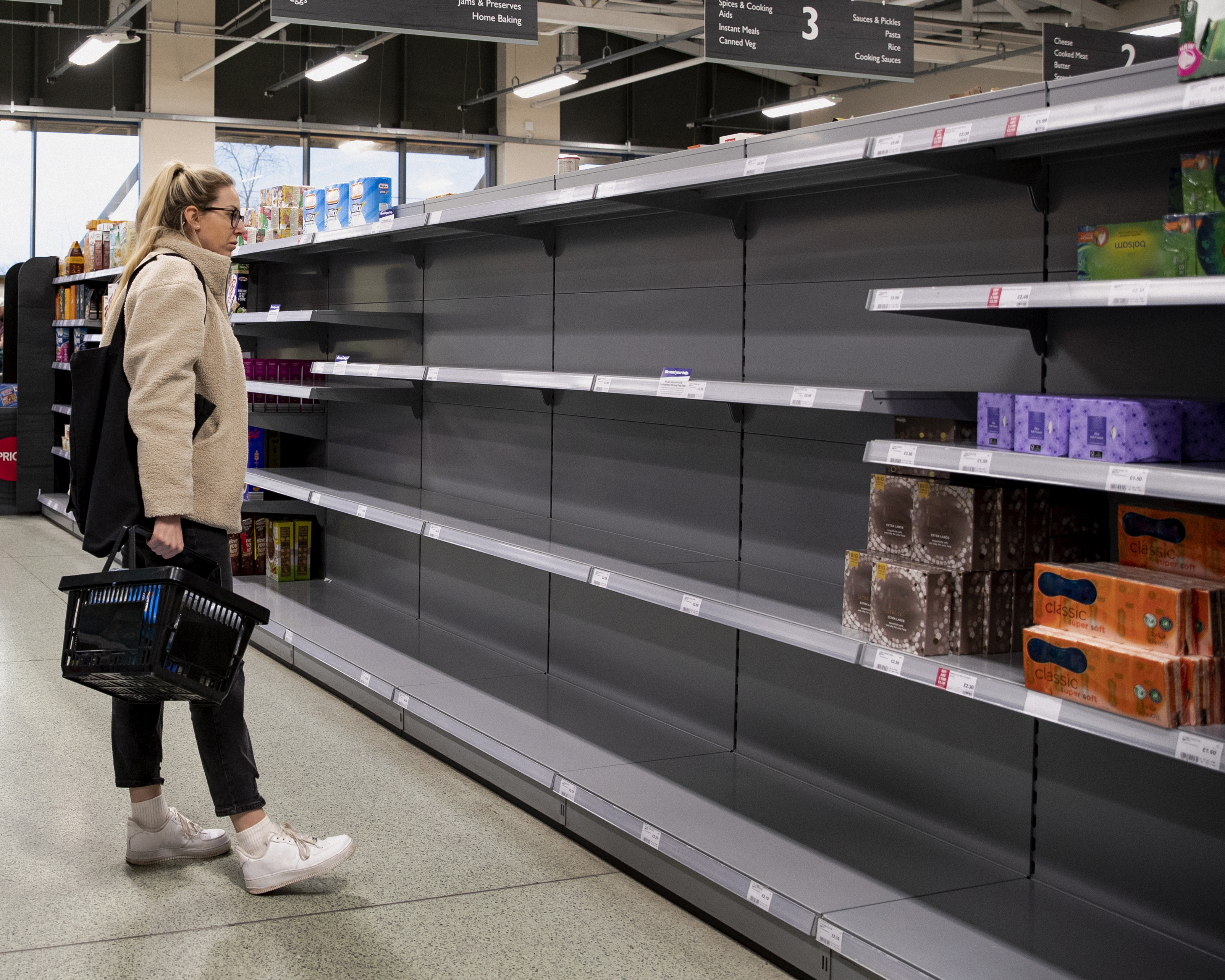 Shopper with a basket glances at mostly empty grocery shelves with few products