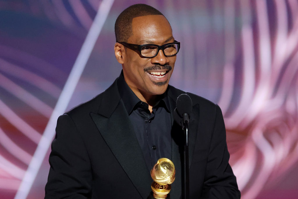 Eddie Murphy holding award, wearing glasses and a black suit, standing at podium with microphone, smiling