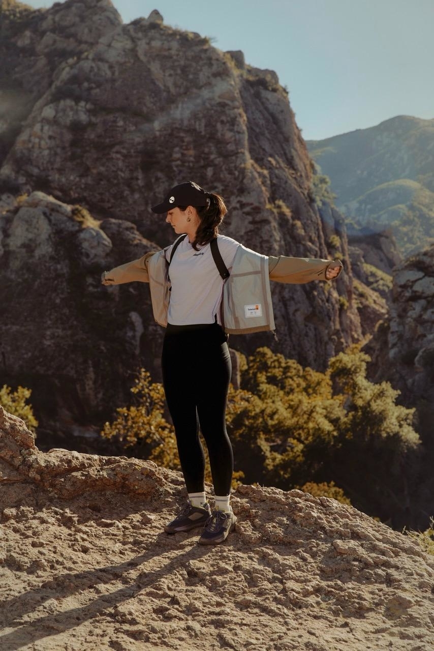 Person in activewear standing with arms outstretched on a rocky outcrop, mountains in the background